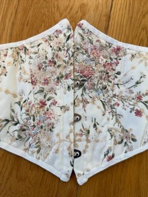 Blommig korsett i pastellfärger - Säljer en romantisk korsett med blommigt mönster i pastelliga toner av rosa, grönt och beige på vit botten. Korsetten har metallknäppning framtill och är kantad med vit söm. Perfekt för att skapa en feminin och trendig look.