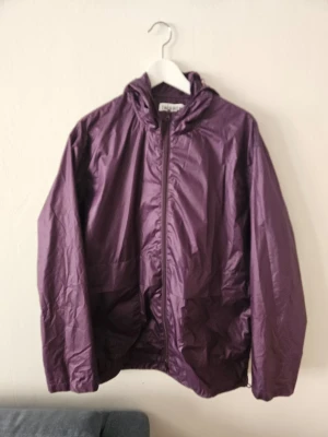 Lila vindjacka - Trés Bien - Snygg lila vindjacka med dragkedja framtill och praktisk huva. Jackan har en lätt och smidig design, perfekt för blåsiga dagar. Tillverkad i ett tunt, glansigt material som ger en sportig look. Passar dig som gillar stilrena och funktionella plagg.