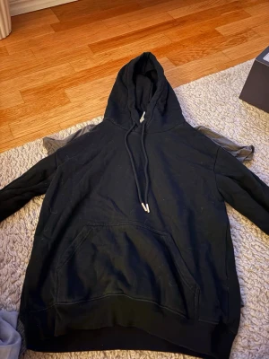 Svart hoodie med ficka och dragsko - En klassisk svart hoodie med stor känguruficka framtill och justerbar dragsko i huvan. Hoodien har en avslappnad passform och är tillverkad i mjukt bomullsmaterial. Perfekt för en chill och stilren look.