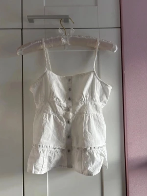 Gullig sommartopp med knappar och justerbar ”rosett” i ryggen - Söt vit linnetopp med smala axelband, knappar framtill och volangdetaljer. Toppen har knyt i ryggen för extra passform och är tillverkad i ett lätt bomullstyg. Storlek xs men skulle själv säga att den sitter som en xxs. Märket ät cotton on. Har själv aldrig använt plagget så det är i utmärkt skick! 