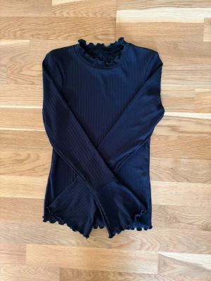 Svart ribbad långärmad halvpolotop - Svart ribbad med långa ärmar och volangdetaljer vid hals, ärmslut och nederkant. Toppens material är stretchigt och mjukt, perfekt för att styla med jeans eller kjol. Enkel men ändå trendig design som passar till många olika looks. Axelbredd 32 cm, längd 54. Aldrig använd. Lapparna bortklippta.