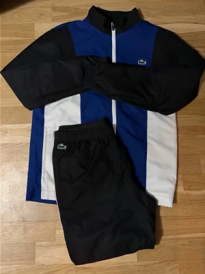 Lacoste träningsset blå svart vit - Snyggt träningsset från Lacoste med jacka och byxor. Jackan har blå, svarta och vita paneler, hög krage och dragkedja framtill. Byxorna är svarta med elastisk midja. Båda plaggen har Lacoste-loggan broderad på bröstet och benet. Perfekt för sport eller chill.