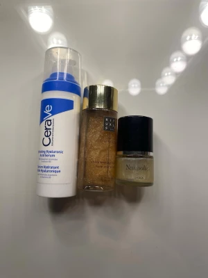 Serum och oljor från CeraVe, Rituals, Nuxe - Tre olika produkter: CeraVe Hyaluronic Acid Serum i vit och blå plastflaska, Rituals Shimmering Body Oil i genomskinlig flaska med guldlock och guldglittrande olja, samt Nuxe Huile Prodigieuse i liten glasflaska med svart lock.