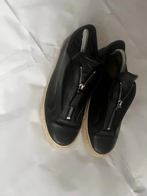 Svarta sneakers från Axel Arigato - Snygga svarta sneakers från Axel Arigato med dragkedja framtill istället för klassiska snören. Ovandelen är i slätt läder och sulan är vit och robust. Minimalistisk design med diskret logga i guld på sidan. Perfekta för dig som gillar stilrena och moderna skor.sulan är bytt och är lite sönder den har gått upp lite där vid dragkedjan men inget som märks!    Pris kan diskuteras!