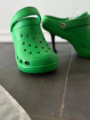 Gröna Crocs-inspirerade pumps - Unika gröna pumps med klassisk Crocs-design och hög smal klack. Skorna har perforerad ovandel, justerbar rem och är tillverkade i lätt plastmaterial. Perfekt för dig som vill sticka ut med en lekfull och trendig stil.