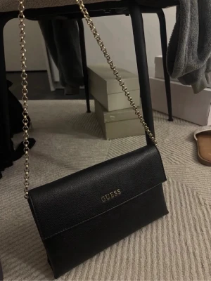 Svart kuvertväska från Guess - Svart axelväska från Guess i slät, strukturerad finish. Smal och stilren modell med lockstängning och guldfärgad kedjerem. Praktisk storlek som rymmer mobil, kort och mindre nödvändigheter. En tidlös väska som funkar både till vardag och kväll.🖤