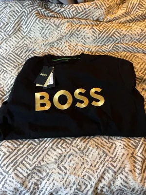 Svart t-shirt från BOSS med guldtryck - Svart tröja från BOSS med stort, glansigt guldtryck över bröstet. Klassisk rund halsringning och korta ärmar. Perfekt för dig som vill sticka ut med en stilren och stilig look. Prisen kan diskuteras 