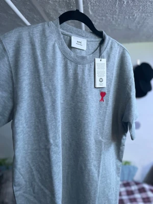 Grå t-shirt från Ami med röd logga - Snygg grå t-shirt från Ami med klassisk passform och rund hals. T-shirten har en liten röd broderad logga på bröstet och är tillverkad i mjuk bomull. Perfekt för en stilren och avslappnad look.