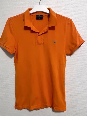 Orange pikétröja från Gant - Snygg orange pikétröja från Gant med klassisk krage och korta ärmar. Tröjan har broderad logga på bröstet och är tillverkad i bomull för en bekväm känsla. Perfekt för en avslappnad och stilren look.