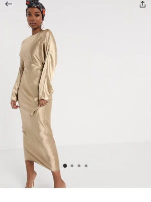 Beige långklänning i satin - Elegant beige långklänning med lång ärm och rund halsringning. Klänningen har en rak, figurnära passform och är tillverkad i ett glansigt satinmaterial som ger en lyxig känsla. Perfekt för dig som vill ha en stilren och sofistikerad look.