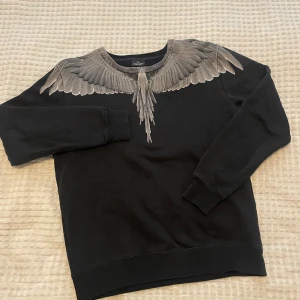 Svart sweatshirt Marcelo Burlon fågel - Svart sweatshirt från Marcelo Burlon med ikoniskt fågeltryck över axlar och bröst. Tröjan är i mjuk bomull och har rund halsringning samt ribbade muddar vid ärmar och nederkant. Perfekt för dig som vill sticka ut med en unik design. Storlek S men passar även M