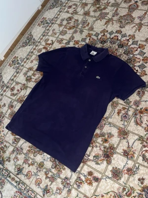 Mörkblå pikétröja från Lacoste - Klassisk mörkblå pikétröja från Lacoste med krage, knappar framtill och den ikoniska krokodilloggan på bröstet. Tröjan har korta ärmar och är tillverkad i mjuk bomull som känns skön mot huden. Perfekt för en clean och sportig stil.