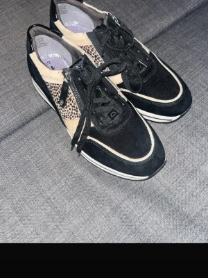 Svarta sneakers med djurmönster - Snygga svarta sneakers från Gabor med beige detaljer och coolt djurmönster på sidorna. Skorna har svarta snören, dragkedja på sidan och vit sula med svart rand. Materialmix av mocka och skinn ger en trendig look.