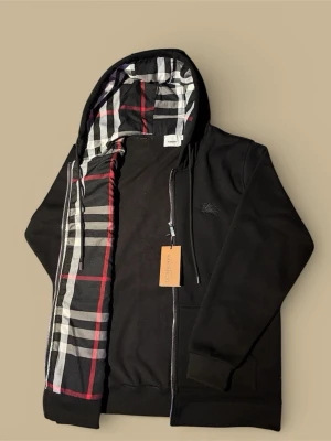 Burberry Kofta - Säljer en svart hoodie från Burberry med klassiskt rutigt foder i huvan och insidan. Tröjan har dragkedja framtill, snörning i huvan och två fickor. Tillverkad i mjuk bomull och har ribbade muddar vid ärmar och nederkant. Perfekt för dig som gillar stilrena detaljer.