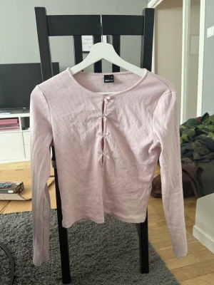 Ljusrosa ribbad topp med knyt - Trendig ljusrosa ribbad topp från Gina Tricot med långa ärmar och tre dekorativa knyt framtill. Toppen har en rund halsringning och är tillverkad i mjuk bomullsblandning. Perfekt för en avslappnad och stilren look.