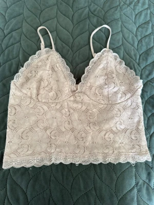 Beige topp/ Bik bok - Säljer en beige topp i spets med blommönster och tunna axelband. Modellen har vacker scallopkant längs både byst och nederkant samt smockad rygg för extra komfort. Perfekt för dig som gillar romantisk och feminin stil.