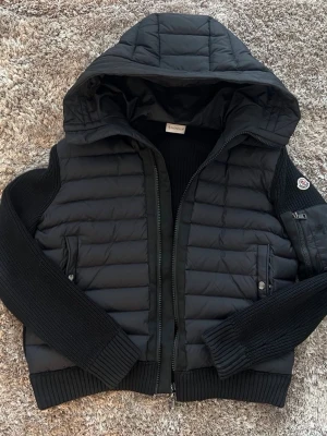 Moncler cardigan - Svart cardiganjacka från Moncler med , vadderad front och stor huva. Ärmar och rygg är ribbstickade i mjukt material. Jackan har dragkedja, två fickor med knapp och Moncler-logga på ärmen. Storlek L men passar M också. Mycket bra skick utan några skador eller defekter förutom en liten sak på bild 4.