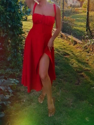 Röd midiklänning med slits - Snygg röd midiklänning med breda axelband och markerad midja. Klänningen har en feminin bystdetalj och en hög slits framtill som ger en cool touch. Perfekt för dig som vill sticka ut. Materialet ser ut att vara ett mjukt tyg med fint fall.