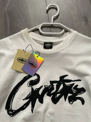 Vit Corteiz t-shirt med grafiskt tryck - Vit t-shirt från Corteiz med stort svart grafiskt tryck framtill och unikt rutmönster med ansikten på ryggen. Klassisk rund hals och korta ärmar. Perfekt för dig som gillar streetwear och vill sticka ut med cool design.