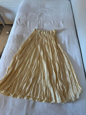 Plisserad beige långkjol i satin - Snygg plisserad långkjol i beige satin med resår i midjan. Kjolen har ett flowigt fall och passar perfekt till både sneakers och boots. Enkel att styla för en trendig look.