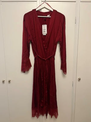 Vinröd morgonrock med spetsdetaljer - Snygg vinröd morgonrock från Gina Tricot i satinliknande material (viskos). Långa ärmar med spetsdetaljer vid ärmslut och nederkant. Knytband i midjan och djup V-ringning. 