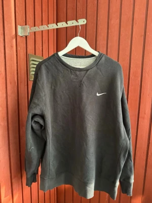 Svart sweatshirt från Nike - En svart sweatshirt från Nike med klassisk rund halsringning och liten vit logga på bröstet. Tröjan har ribbade muddar vid ärmslut och nederkant. Perfekt för en avslappnad och sportig stil.