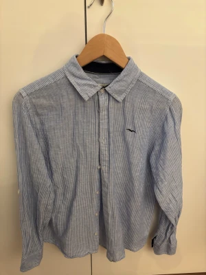 Blårandig skjorta från hamilton republic - Klassisk blårandig skjorta från hamilton republic med vit knappstängning och liten broderad logga på bröstet. Skjortan har lång ärm, krage och är tillverkad i ett tunt, skönt bomullstyg. Perfekt för en stilren och avslappnad look.