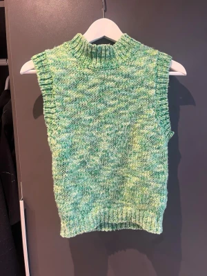 Grön stickad väst från NA-KD - Säljer en grön stickad väst från NA-KD med hög krage och ribbade kanter. Västen har en melerad look i olika gröna nyanser och är perfekt att styla över en skjorta eller t-shirt för en trendig lager-på-lager-look.