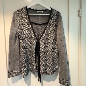 Svartvit mönstrad kofta med knytband från Odd Molly - Snygg svartvit kofta med grafiskt mönster och långa ärmar. Djup V-ringning med svart kant och knytband framtill. Mjuk och tunn stickning, perfekt för lager-på-lager. Unik design med detaljerade mönster både fram och bak. Odd Molly storlek 1.