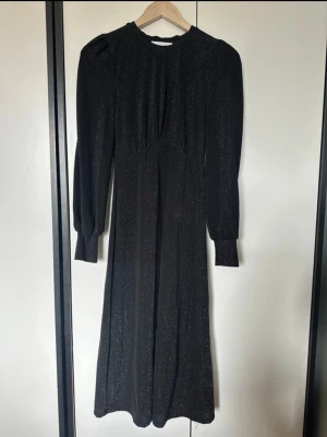 Svart glittrig långklänning - En svart långklänning med glittrigt tyg och långa ärmar. Klänningen har en rund halsringning och markerad midja, vilket ger en snygg siluett. Perfekt för dig som vill sticka ut med en elegant och trendig look.