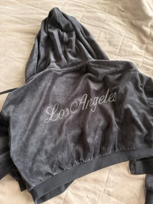 Mörkgrå velourhoodie  - Snygg mörkgrå hoodie i mjuk velour med dragkedja och huva. På ryggen står det 'Los Angeles' i glittriga stenar och framtill finns en liten stjärna broderad. Perfekt för en chill och trendig look.