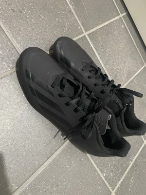 Svarta Adidas fotbollsskor - Säljer ett par helsvarta fotbollsskor från Adidas med klassisk design och snörning. Skorna har syntetiskt material och mönstrad sula för bra grepp på planen. Diskreta svarta ränder på sidorna och låg profil. Perfekta för dig som vill ha stilrena skor till match eller träning.