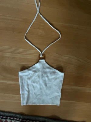 Vit ribbad halterneck topp - Snygg vit ribbad topp med halterneck och cool detalj i form av en brun plastspänne framtill. Toppen är ärmlös och croppad, perfekt till jeans eller kjol. Enkel och stilren design som passar till många olika looks.