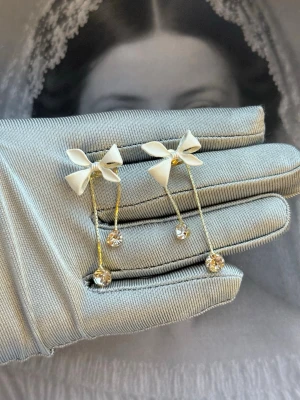 Guldiga örhängen med rosett och strass - Snygga örhängen med tunn guldfärgad båge, vita rosetter upptill och glittrande strassdetaljer längst ut. Perfekt för dig som gillar en söt och trendig look. Materialet är metall och plast, och designen är lekfull men elegant.