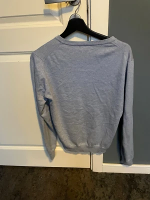 Ljusgrå V-ringad tröja Lyle & Scott - Snygg ljusgrå tröja från Lyle & Scott med klassisk V-ringning och den ikoniska gula örnloggan på bröstet. Tillverkad i mjukt material och har långa ärmar samt ribbade muddar vid ärmslut och nederkant. Perfekt för en clean och stilren look.