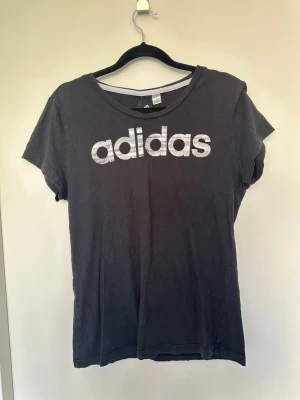 Svart Adidas t-shirt med logga Medium - Snygg svart t-shirt från Adidas med stor vit logga framtill. Klassisk rund halsringning och korta ärmar. Tillverkad i mjuk bomull som känns skön mot huden. Perfekt för en sportig och avslappnad stil.