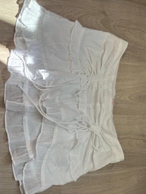 Vit kortkjol med volanger och snörning - Söt vit kortkjol med volangdetaljer och snörning framtill. Kjolen har flera lager och en luftig känsla, perfekt för varma dagar. Tillverkad i ett lätt material som ger en flowy look. Passar dig som gillar romantisk och trendig stil. Med prislapp kvar
