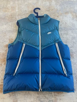 Blå dunväst från Nike - Säljer en stilren dunväst från Nike i två nyanser av blått med vita dragkedjor som ger ett snyggt och sportigt uttryck. Västen har hög krage, två praktiska sidofickor med dragkedja samt Nike-logga på bröstet.  Perfekt som ett lager-på-lager-plagg och passar utmärkt till både vardags- och streetwear-stilar.  Västen är i storlek L och har endast använts vid fyra tillfällen. Skicket är därför mycket fint, i princip som nytt.  Pris kan diskuteras vid seriöst intresse.