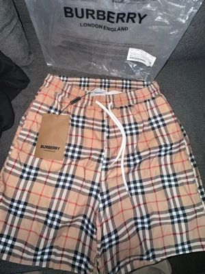 Beige rutiga shorts från Burberry - Säljer ett par klassiska beige badshorts/shorts från Burberry med det ikoniska rutiga mönstret i svart, vitt och rött. Shortsen har elastisk midja med vit snörning och är perfekta för en trendig och avslappnad stil. Kommer med originalpåse och tag.