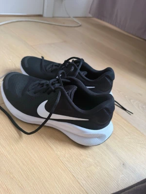 Svarta Nike sneakers med vit sula - Snygga svarta Nike sneakers med vit sula och vit swoosh på sidan. Skorna har en låg profil och är tillverkade i meshmaterial för bra ventilation. Perfekta för dig som vill ha en clean och sportig look.