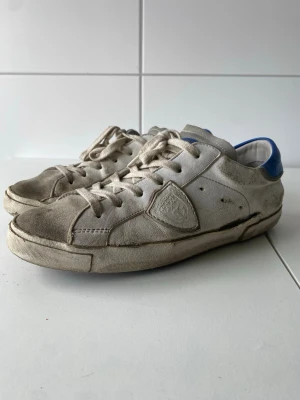 Philippe Model vita sneakers med blå detalj - Säljer eller byter ett par vita Philippe Model sneakers med blå hälkappa och klassisk låg siluett. Skorna har läder- och mockadetaljer, vita snören och en ikonisk sköldlogga på sidan. Perfekta för dig som gillar en avslappnad och trendig stil.