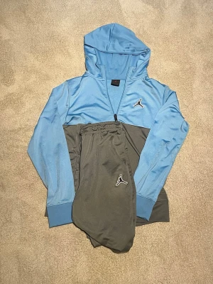 Blå och grå hoodie från Jordan - Snygg hoodie från Jordan i ljusblått och grått med dragkedja och huva. Ikoniska Jumpman-loggan broderad på bröstet. Tillverkad i mjukt och stretchigt material som känns skönt mot huden. Perfekt för dig som gillar sportig och avslappnad stil.
