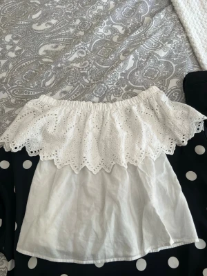 Vit offshoulder topp Gina Tricot - Supersöt vit offshoulder topp från Gina Tricot med broderad spetsvolang runt axlarna. Toppen är ärmlös och har en luftig passform, perfekt för varma dagar. Tillverkad i bomull för en skön känsla.