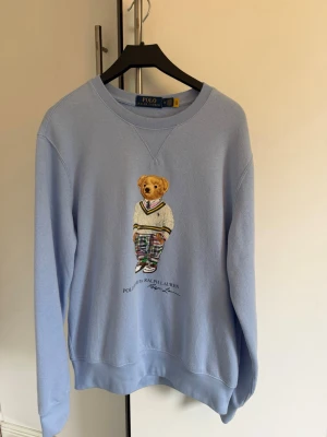 Polo Bear Ralph Lauren Ljusblå Tröja - Stl M - En ljusblå sweatshirt med rund halsringning. Tröjan har ett tryck av Polo Bear by Ralph Lauren på framsidan. Mycket fin tröja, använd max 10 gånger.   Ett väldigt litet hål på framsidan, se bild 4. Knappt synligt och det går troligen att sy igen väldigt enkelt. 