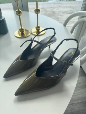 Olivgröna lackpumps från Zara - Säljer ett par olivgröna pumps från Zara i glansigt lackmaterial. Skorna har spetsig tå, smal klack och justerbar rem runt ankeln. Perfekta för dig som vill ha en trendig och elegant look med en unik färg. Helt nya! Endast testade 