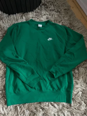 Grön crewneck sweatshirt från Nike - Snygg grön sweatshirt från Nike med klassisk vit broderad logga på bröstet. Tröjan har rund halsringning, långa ärmar och ribbade muddar vid ärmslut och nederkant. Perfekt för en avslappnad och sportig stil.