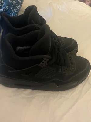 Svarta Air Jordan 4 sneakers - Säljer ett par svarta Air Jordan 4 sneakers med klassisk Jumpman-logo på hälen. Skorna har en hög siluett, meshpaneler och detaljer i läder och syntet. Perfekt för dig som vill ha en ikonisk och stilren sneaker med cool design.
