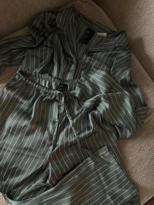 Grön randig pyjamas från H&M - Snygg grön pyjamas från H&M i mjukt, glansigt material med vita tunna ränder. Setet består av en långärmad skjorta med krage och knappar samt matchande byxor med resår i midjan. Perfekt för chill hemma eller mysiga kvällar. Endast testad