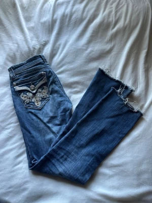 Vintage jeans - Säljer ett par blå jeans från Grace in LA med snyggt broderad bakficka och slitna, råa benslut. Jeansen har låg midja, klassisk femficksmodell och coola detaljer med silverknappar och dekorationer. Perfekta för en avslappnad och trendig look.