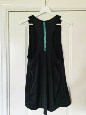 Svart sporttopp från H&M - Svart träningslinne från H&M med racerback och en turkos detalj på ryggen. Toppen är ärmlös och har en loose passform, perfekt för träning eller avslappnade dagar. Materialet är mjukt och lätt polyester.